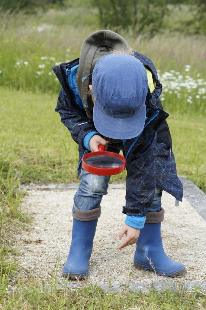 Child in blue hat examines ground with red magnifying glass, wearing boots and jacket, in a grassy field.
