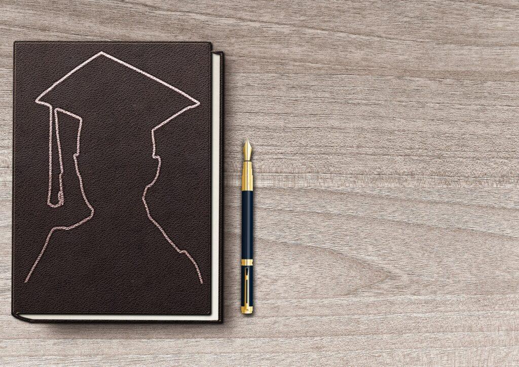Brown journal with a graduation cap outline beside a fountain pen on a wooden surface.