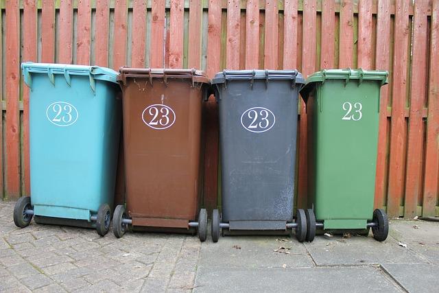 Four wheeled bins of different colors numbered 23 lined up against a wooden fence.