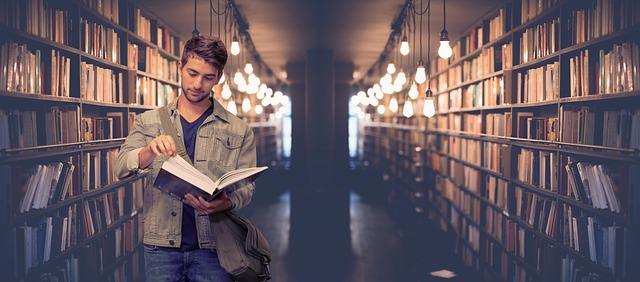 Man reading a book in a dimly lit library aisle, surrounded by shelves of books.